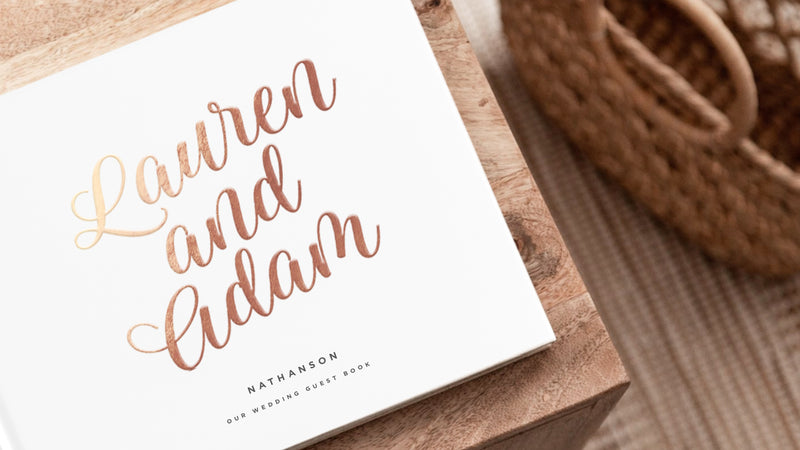 white hard cover guest book sitting on wood table with gold lettering that reads "Lauren and Adam"